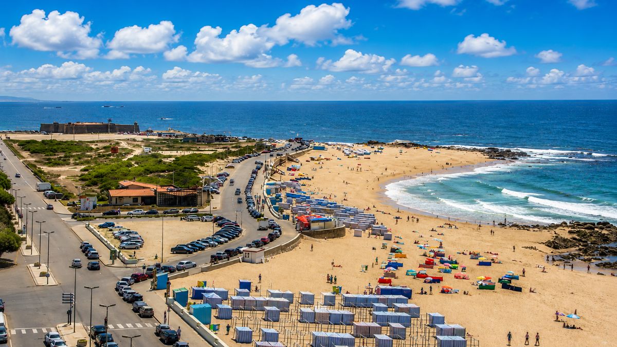 Fotografia aérea da Praia do Turismo em Vila do Conde mostrando uma extensa faixa de areia dourada com guarda-sóis coloridos e barracas listradas azuis e brancas. À esquerda há uma avenida marginal curvada repleta de carros estacionados e em movimento. Ao fundo o Oceano Atlântico azul sob um céu ensolarado com nuvens brancas e a antiga fortaleza de pedra na foz do rio.