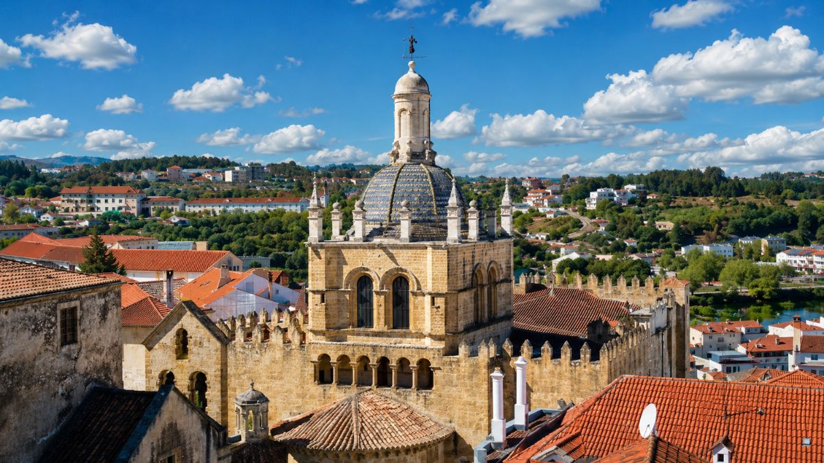 Fachada de pedra e torre lanterna da Sé Velha de Coimbra com telhados laranjas e rio Mondego ao fundo sob céu azul.