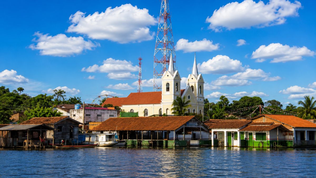 Uma imagem panorâmica de uma comunidade ribeirinha na Amazônia, mostrando casas flutuantes e palafitas de madeira ancoradas na margem do rio, com uma igreja católica amarela de duas torres sineiras no centro e uma alta torre de comunicação treliçada vermelha e branca atrás dela, sob um céu azul ensolarado com nuvens brancas fofas.