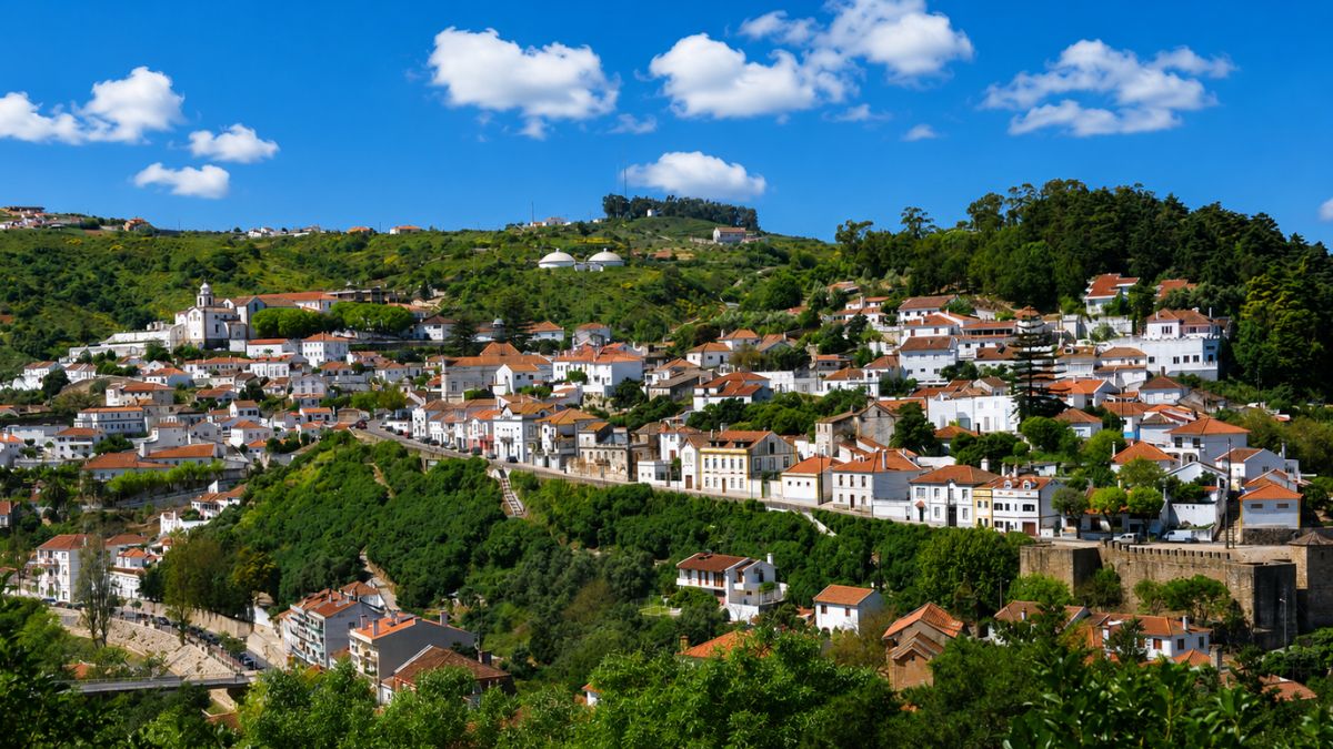 Vista panorâmica de Alenquer com casas brancas dispostas em anfiteatro na encosta verde sob céu azul com nuvens brancas.