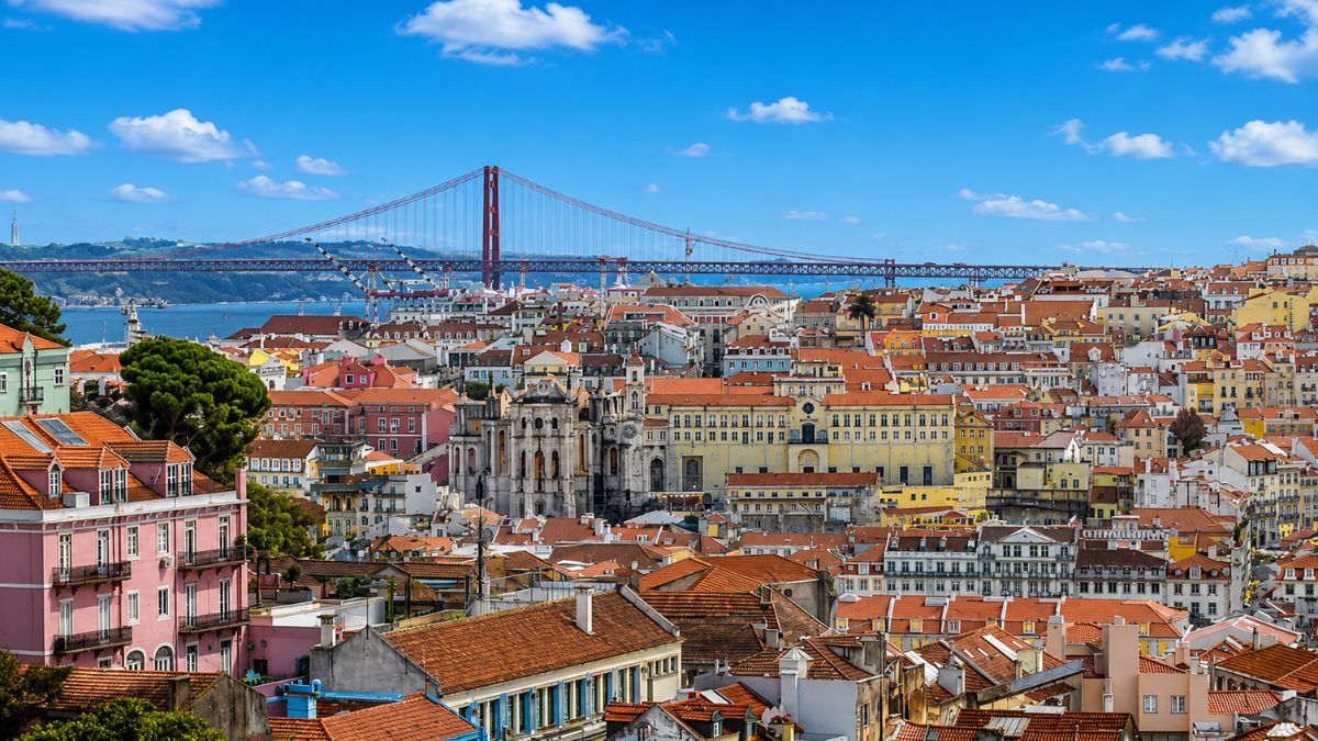 Fotografia colorida com vista aérea de Lisboa exibindo o casario antigo de telhados laranjas, monumentos históricos, o Rio Tejo ao fundo e a grande ponte suspensa vermelha sob um céu azul com nuvens.