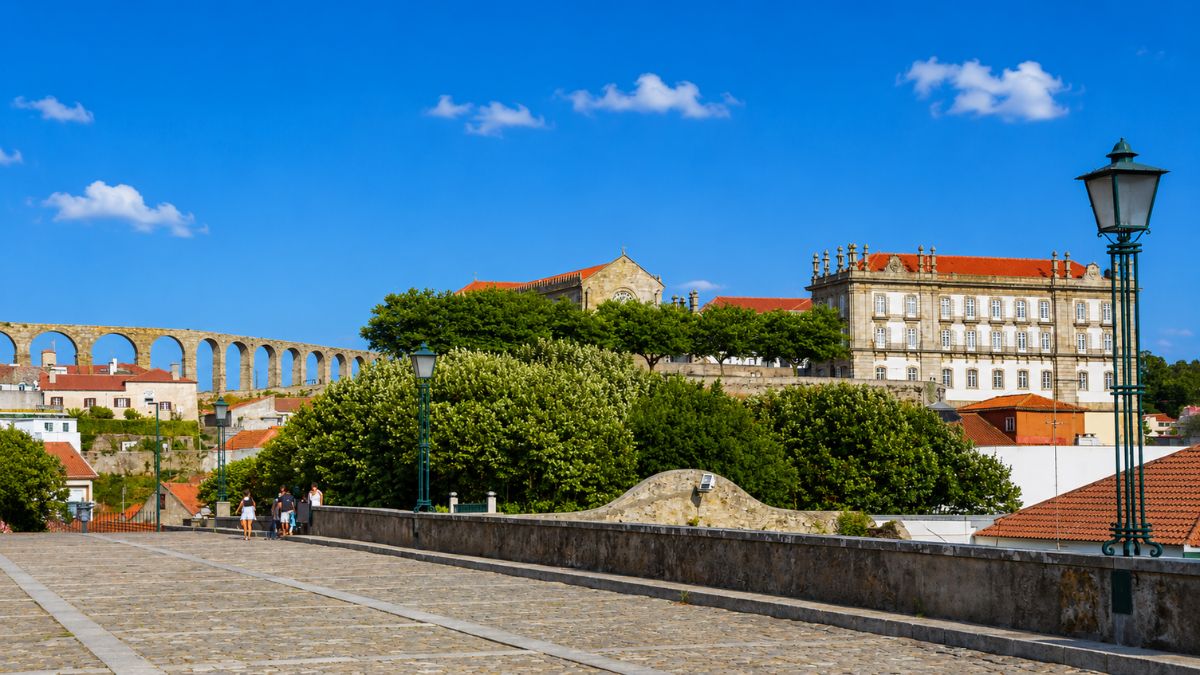 Fotografia colorida tirada de uma ponte de pedra em Vila do Conde mostrando ao fundo o imponente Mosteiro de Santa Clara à direita e os arcos de pedra do Aqueduto de Santa Clara estendendo-se para a esquerda sob um céu azul limpo com poucas nuvens.