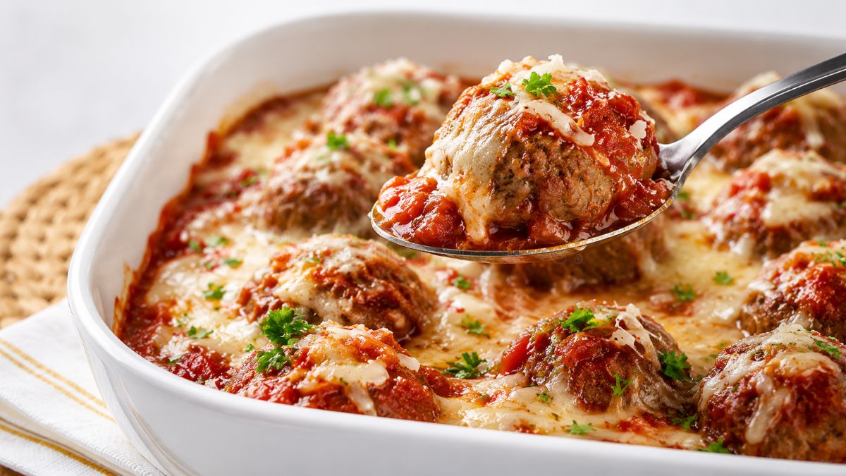 Almôndegas de peru assadas e recheadas com queijo mozarela derretido cobertas por molho de tomate artesanal e parmesão em uma travessa branca com uma colher de metal servindo uma unidade.