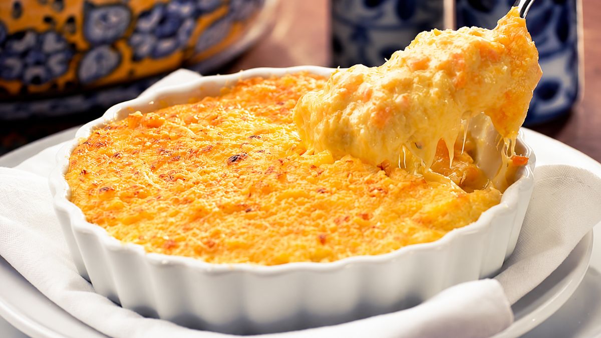 Um ramekin branco com bacalhau espiritual gratinado, uma colher de sopa tirando uma porção com queijo derretido e cenoura ralada.