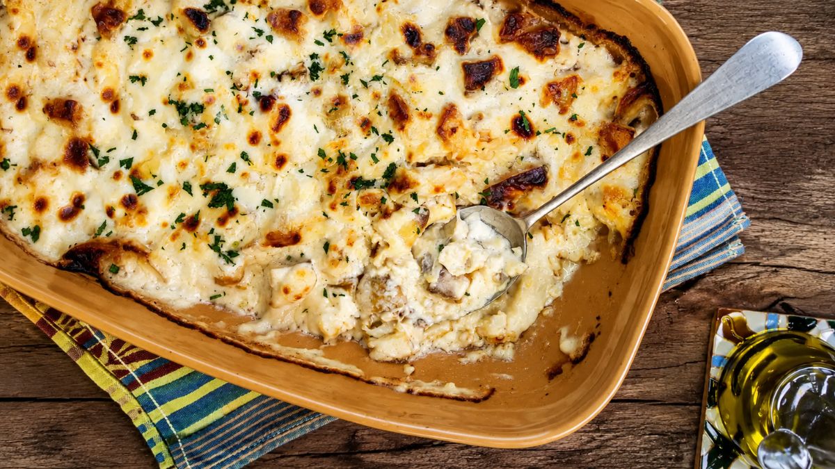 Bacalhau com natas português servido em uma travessa de cerâmica cerimonial com uma colher de metal retirando uma porção cremosa do prato gratinado com queijo dourado e salpicado com ervas frescas sobre uma mesa rústica de madeira.