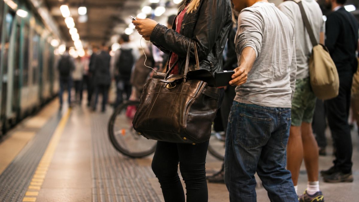 Homem de camiseta cinza retirando sorrateiramente uma carteira escura de dentro da bolsa aberta de uma mulher distraída no celular na plataforma do metrô de Porto.