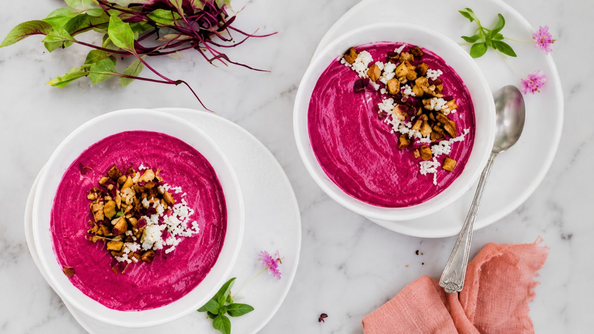 Vista superior de duas tigelas brancas com creme de beterraba roxo vibrante decorado com croutons crocantes, queijo ralado e brotos, posicionadas sobre uma mesa de mármore claro com uma colher de metal e folhas verdes ao fundo.