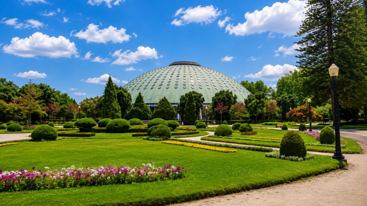 Grande cúpula verde do Pavilhão Rosa Mota cercada por jardins gramados com flores e árvores sob céu azul ensolarado.