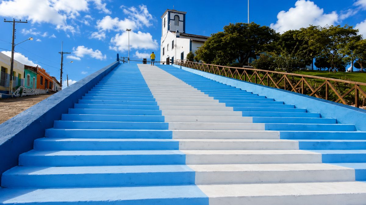 Escadaria monumental pintada com listras azuis e brancas que leva até a Igreja Matriz de Nossa Senhora da Conceição em Monte Santo, Bahia, sob céu azul com nuvens.