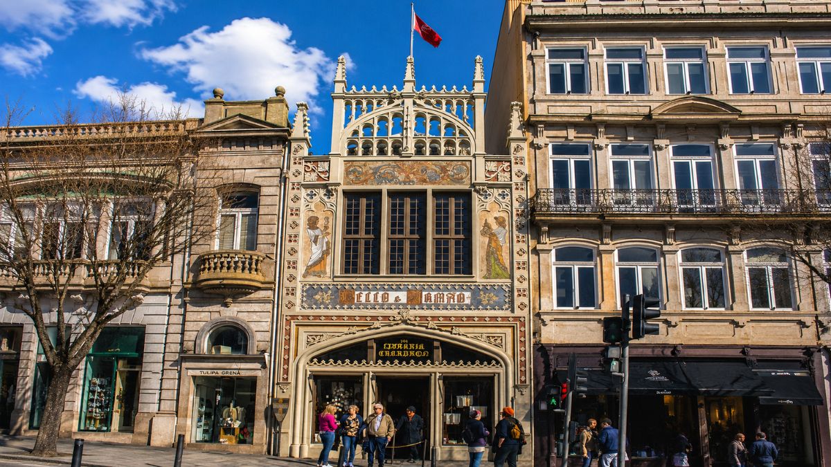 Fachada da Livraria Lello em Porto com arquitetura neogótica detalhada, janelas grandes, pinturas artísticas e pedestres na calçada em dia ensolarado.