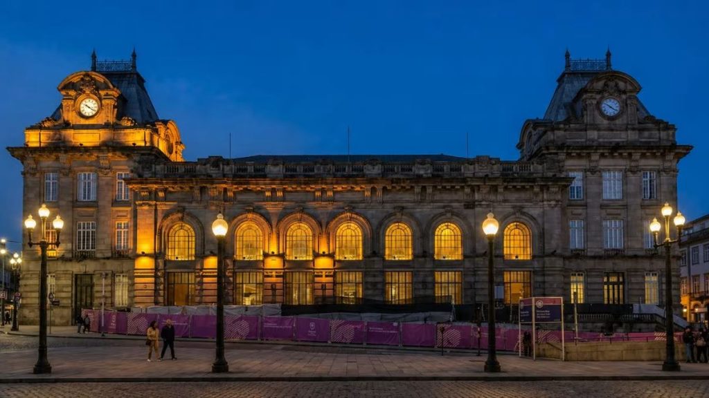 Fachada iluminada da Estação de São Bento no Porto à noite com arquitetura clássica, grandes janelas em arco acesas e postes de luz.