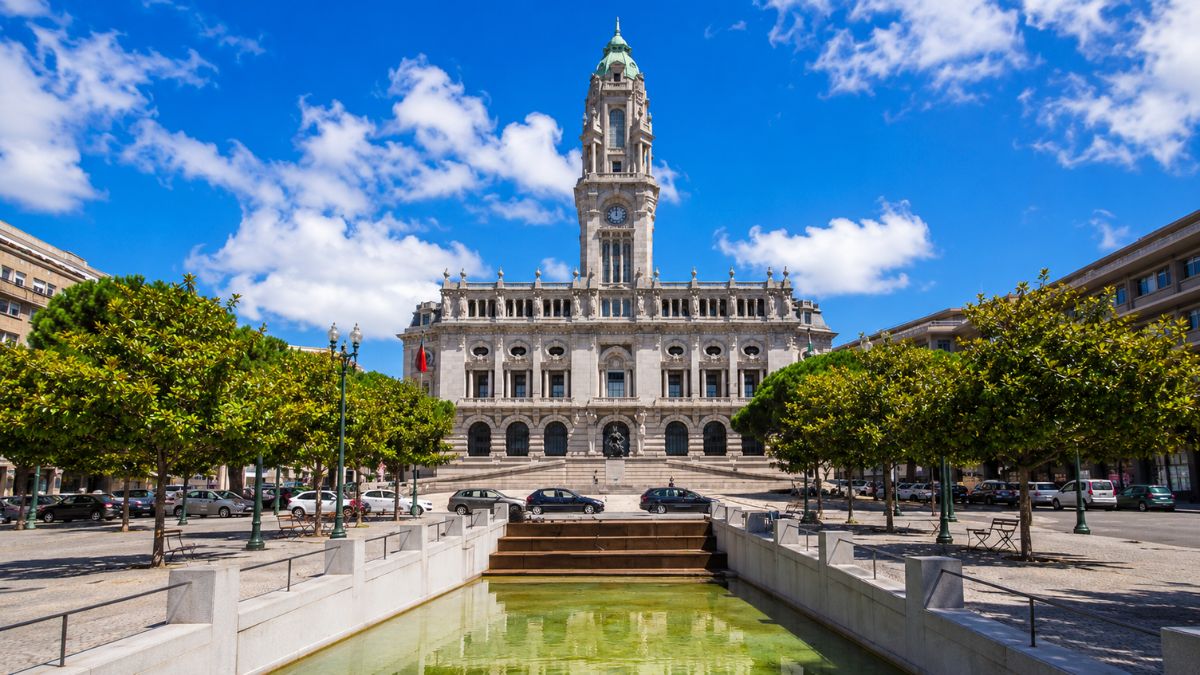 Fachada principal do edifício da Câmara Municipal do Porto vista a partir da Praça da Liberdade com espelho d'água em primeiro plano sob céu azul.