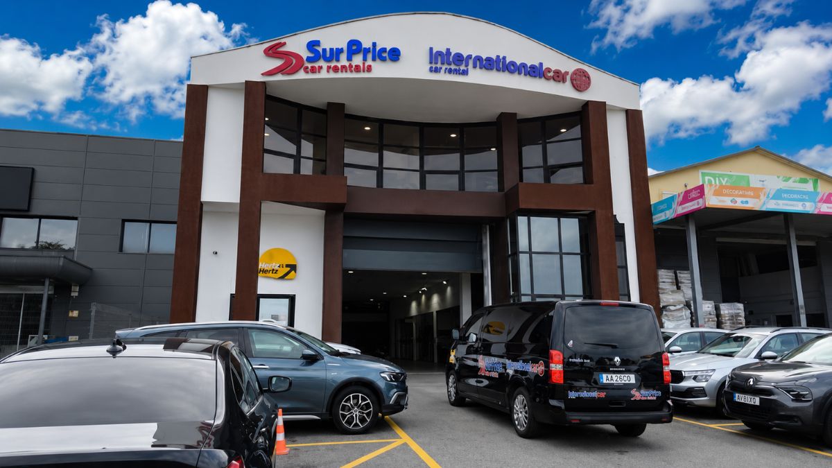 Fachada de prédio comercial moderno com letreiros das locadoras SurPrice Car Rentals e International Car Rental com carros estacionados à frente sob céu azul.