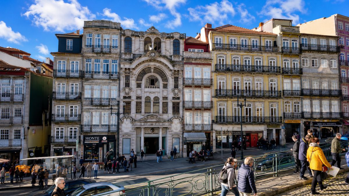 Prédios antigos com fachadas detalhadas em pedra e azulejos coloridos na cidade do Porto, com pessoas caminhando em uma rua movimentada sob céu azul com nuvens.