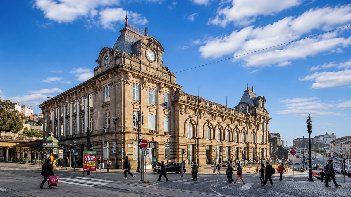 Fachada externa em pedra da Estação de São Bento com sua arquitetura detalhada e relógio central sob céu azul com pessoas caminhando na calçada.