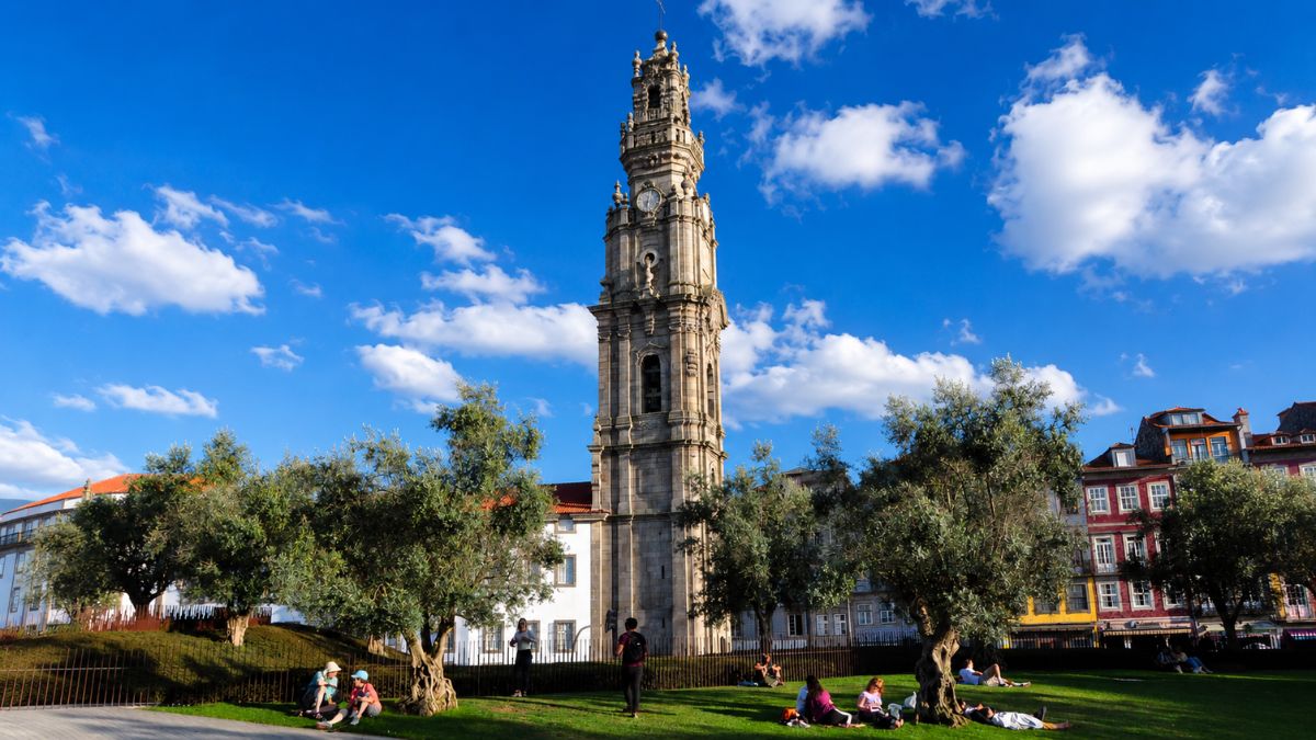 Torre dos Clérigos em estilo barroco sob céu azul com nuvens brancas vista a partir de um gramado arborizado com pessoas descansando no centro do Porto.