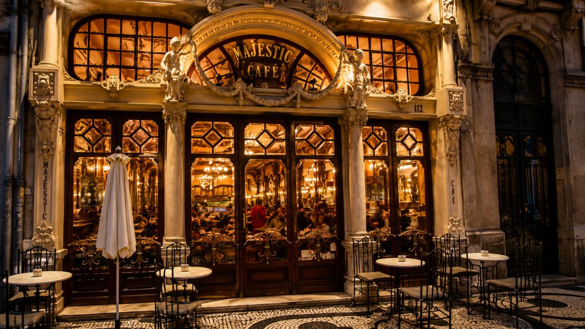 Fachada iluminada do Café Majestic exibindo esculturas de querubins, grandes vitrines de madeira e mesas na calçada de pedra portuguesa.