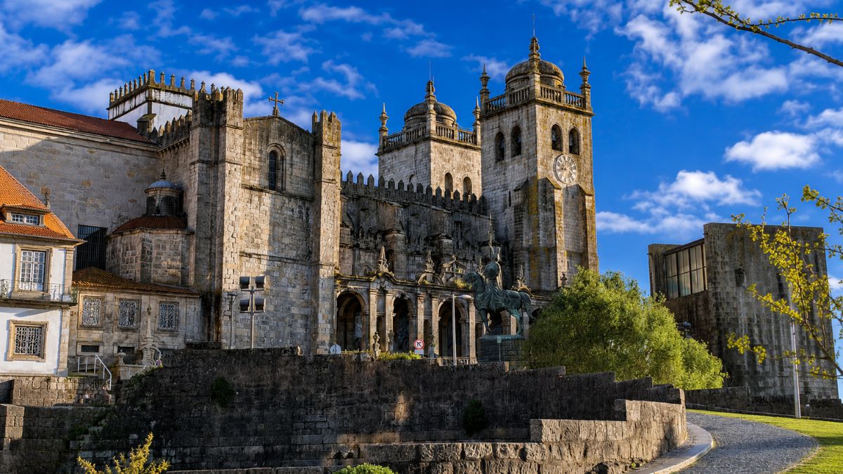 Vista exterior da fachada lateral em granito da Catedral da Sé do Porto, destacando a galilé barroca de Nicolau Nasoni, as duas torres sineiras simétricas com cúpulas bulbosas sob céu azul e a estátua equestre do guerreiro Vímara Peres em primeiro plano.