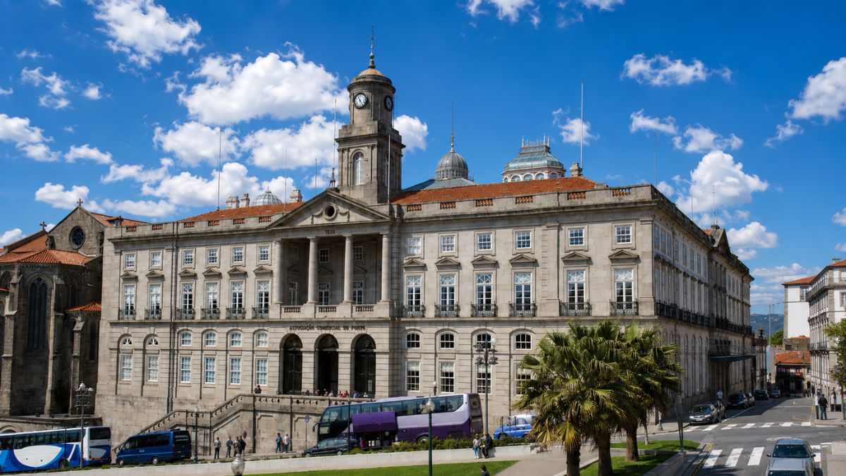 Fachada externa em pedra do Palácio da Bolsa com sua torre do relógio central cercada por gramado, palmeiras e carros sob céu azul.