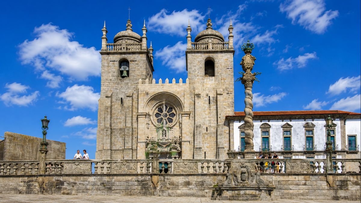 Vista frontal exterior da fachada românica em granito da Catedral da Sé do Porto, destacando a rosácea central do século XII, as duas torres sineiras com cúpulas barrocas, a balaustrada de pedra e o pelourinho monumental em primeiro plano sob céu azul.
