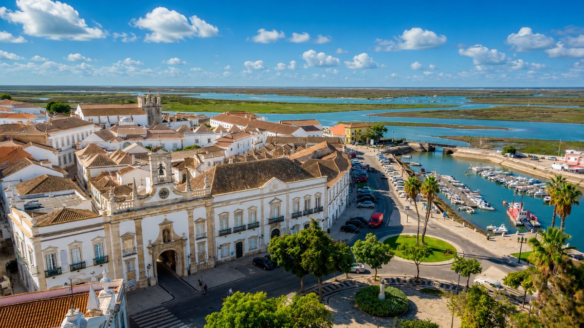 Vista aérea da Cidade Velha de Faro com o Arco da Vila, carros estacionados na Marina, barcos ancorados e canais da Ria Formosa ao fundo sob céu azul.