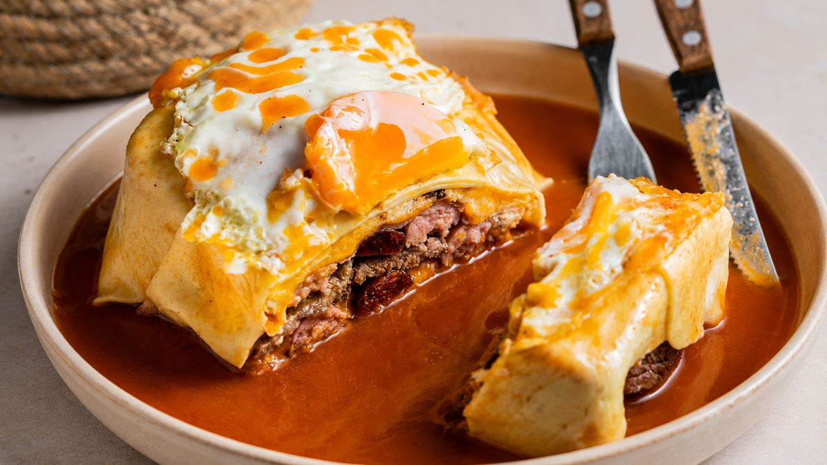 Fotografia em plano detalhe de uma francesinha especial cortada ao meio em um prato profundo com molho denso, revelando as camadas de bife de carne de vaca, embutidos e queijo derretido, com um ovo estrelado de gema mole escorrendo no topo.