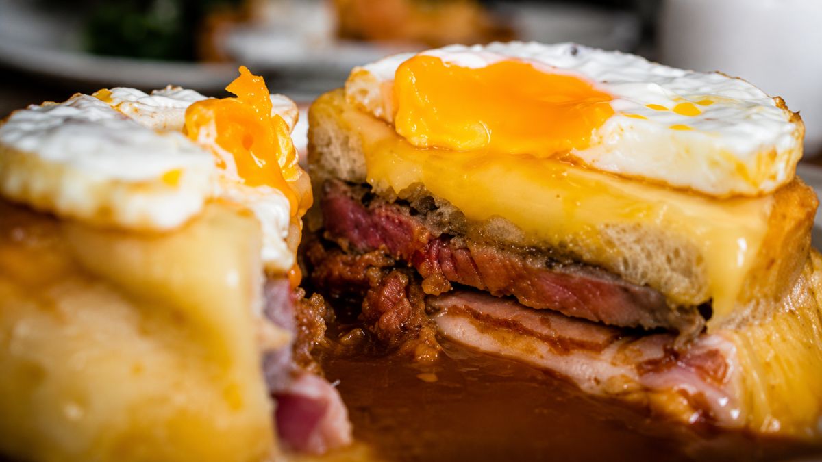 Sanduíche francesinha especial cortado ao meio mostrando camadas de bife, presunto e embutidos sob queijo derretido, ovo frito e molho picante.