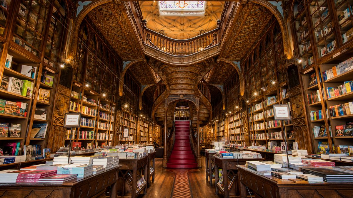 Interior da Livraria Lello com estantes de madeira repletas de livros e a famosa escadaria sinuosa com degraus vermelhos no centro sob teto decorado.