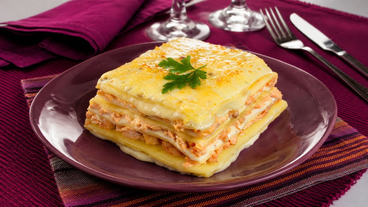 Uma porção quadrada de lasanha de salmão e espinafres com queijo gratinado e uma folha de salsa no topo servida em um prato roxo.