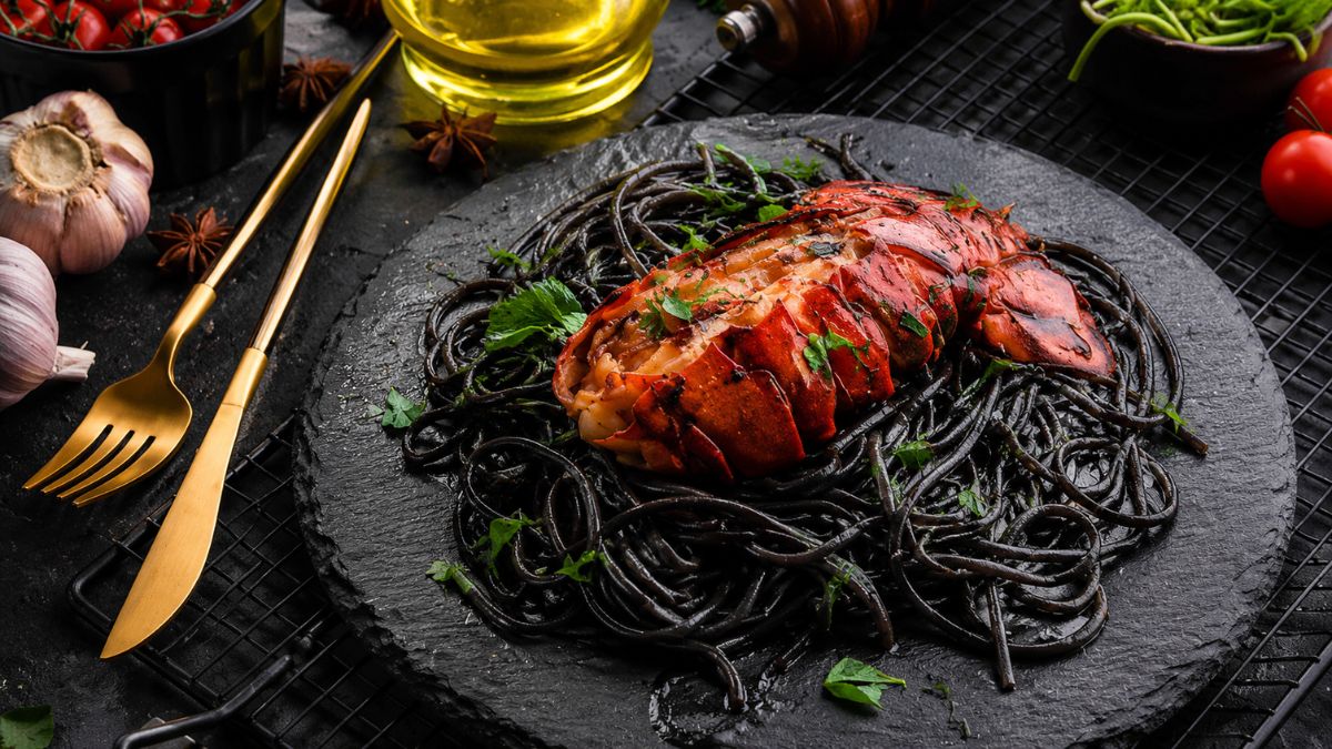 Prato gourmet de espaguete preto com tinta de lula servido com uma cauda de lagosta grelhada bem dourada por cima, decorado com ervas frescas e posicionado ao lado de talheres dourados em um fundo escuro.