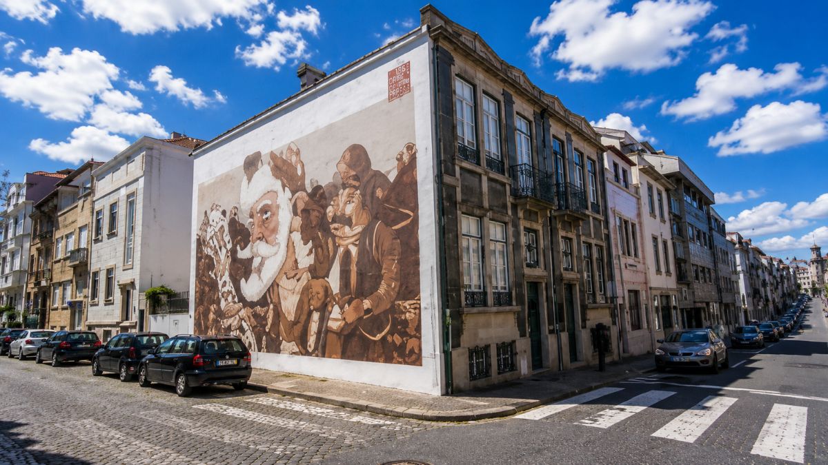 Grande pintura mural em tons de marrom na parede lateral de um prédio residencial em uma esquina com carros estacionados sob céu azul.