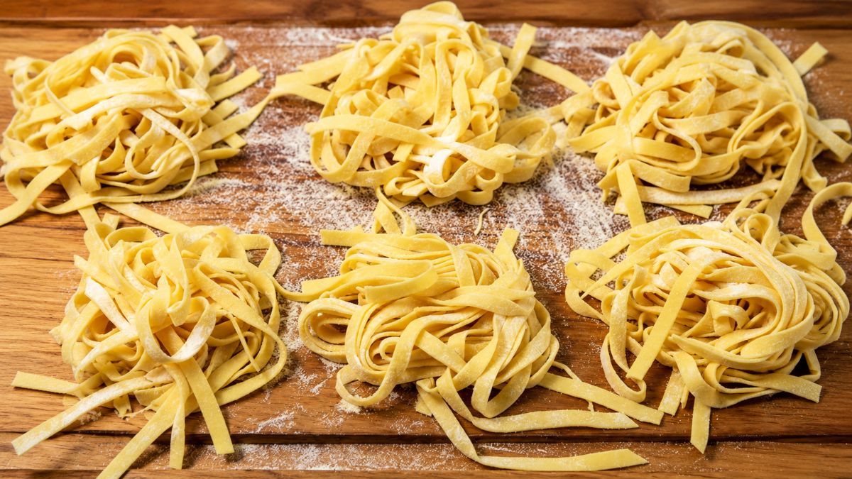 Seis ninhos de massa fresca artesanal tipo fettuccine dispostos em duas fileiras sobre uma tábua de madeira polvilhada com farinha, em plano fechado.