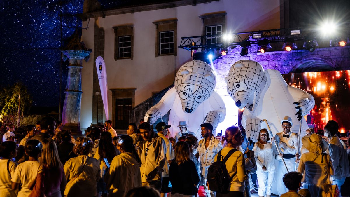 Artistas vestidos de branco conduzem duas grandes figuras insufláveis de ursos brancos iluminados no centro histórico durante a noite branca em Vila do Conde, rodeados por uma multidão de visitantes sob um céu estrelado.