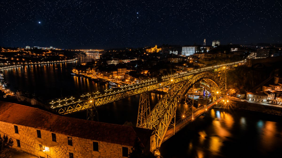 Vista aérea noturna da Ponte Dom Luís I iluminada sobre o Rio Douro ligando Porto a Vila Nova de Gaia sob um céu estrelado.