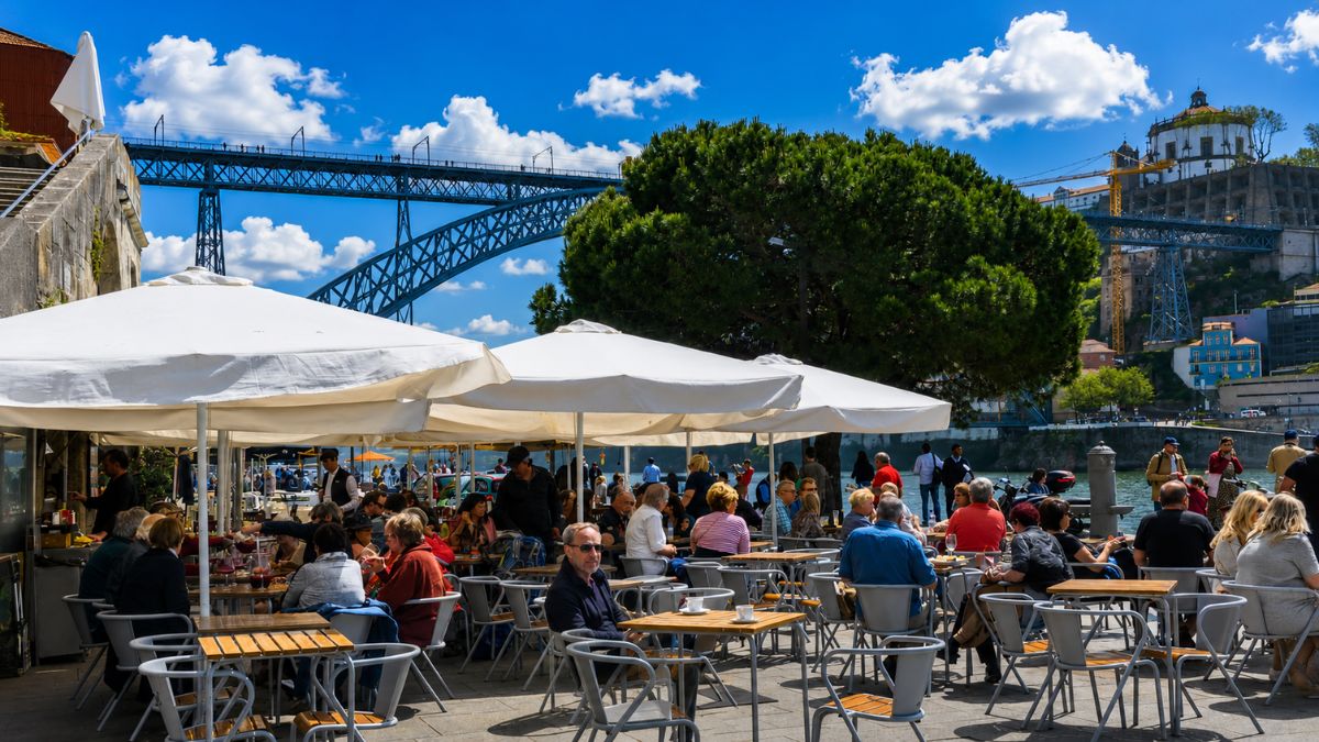 Restaurante com mesas ao ar livre e guarda-sóis brancos repleto de clientes almoçando na Ribeira do Porto com vista para o Rio Douro e a Ponte Dom Luís I sob céu azul.