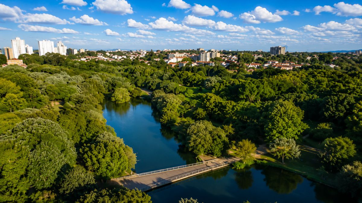 Vista aérea do Parque da Cidade do Porto mostrando grande lago com ponte cercado por intensa mata verde e edifícios ao fundo