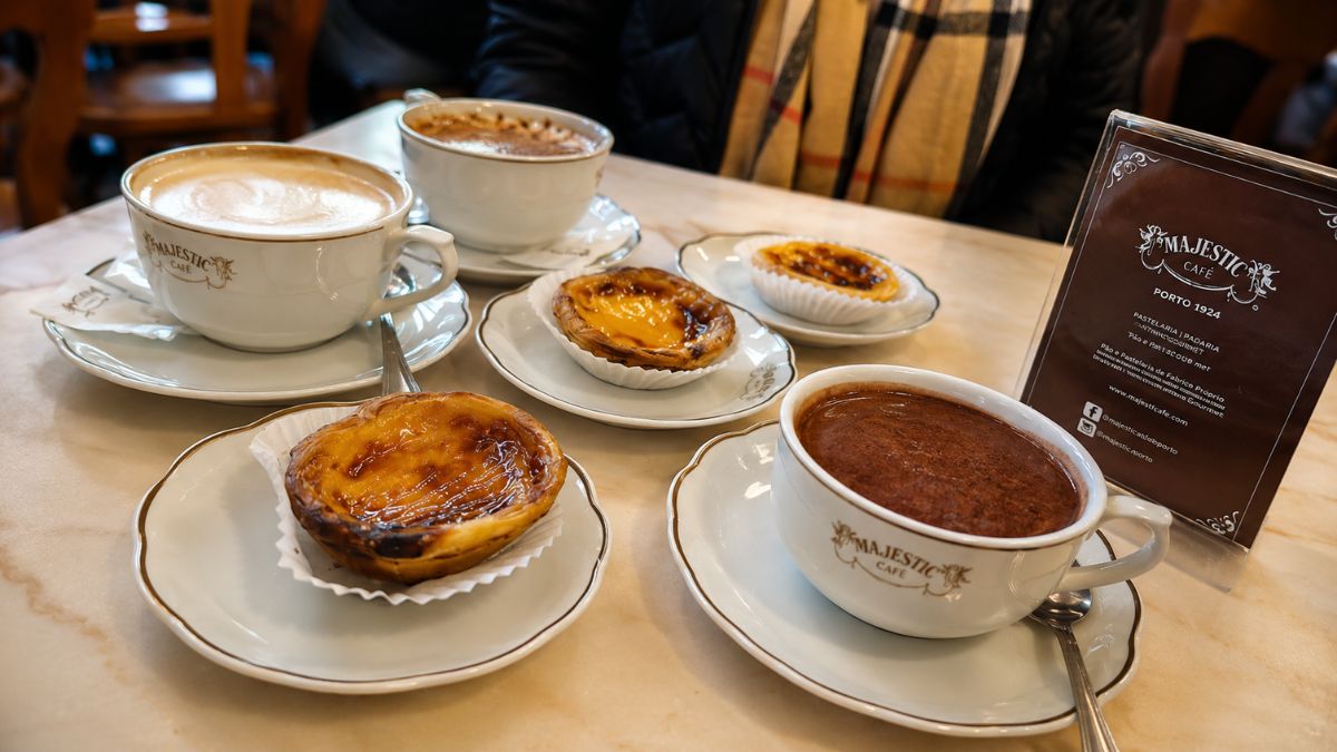 Mesa de mármore no Majestic Café com pastéis de nata dourados em pires e xícaras de café com leite e chocolate quente.