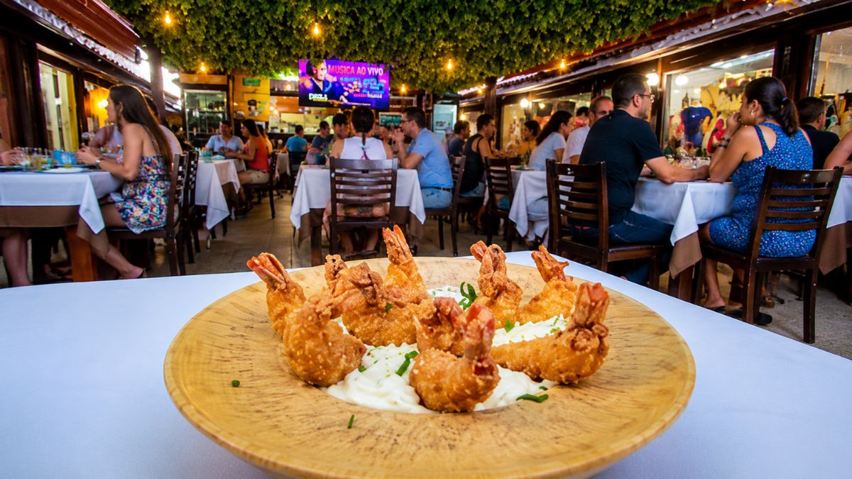 Prato de camarão empanado frito disposto em formato circular sobre molho branco em uma travessa de madeira com o salão de um restaurante movimentado e iluminado ao fundo.