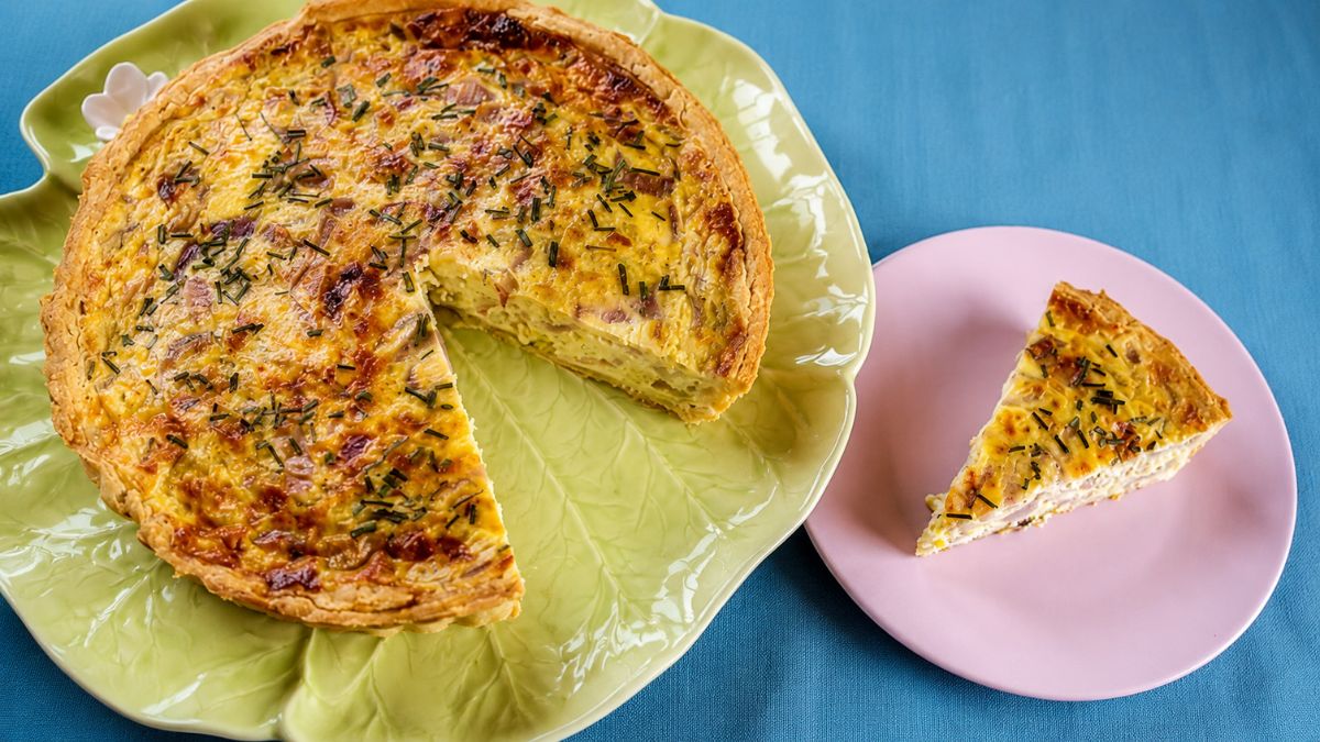 Quiche de alho francês e cebola roxa com massa quebrada dourada em prato verde ao lado de uma fatia servida em prato rosa sobre fundo azul.