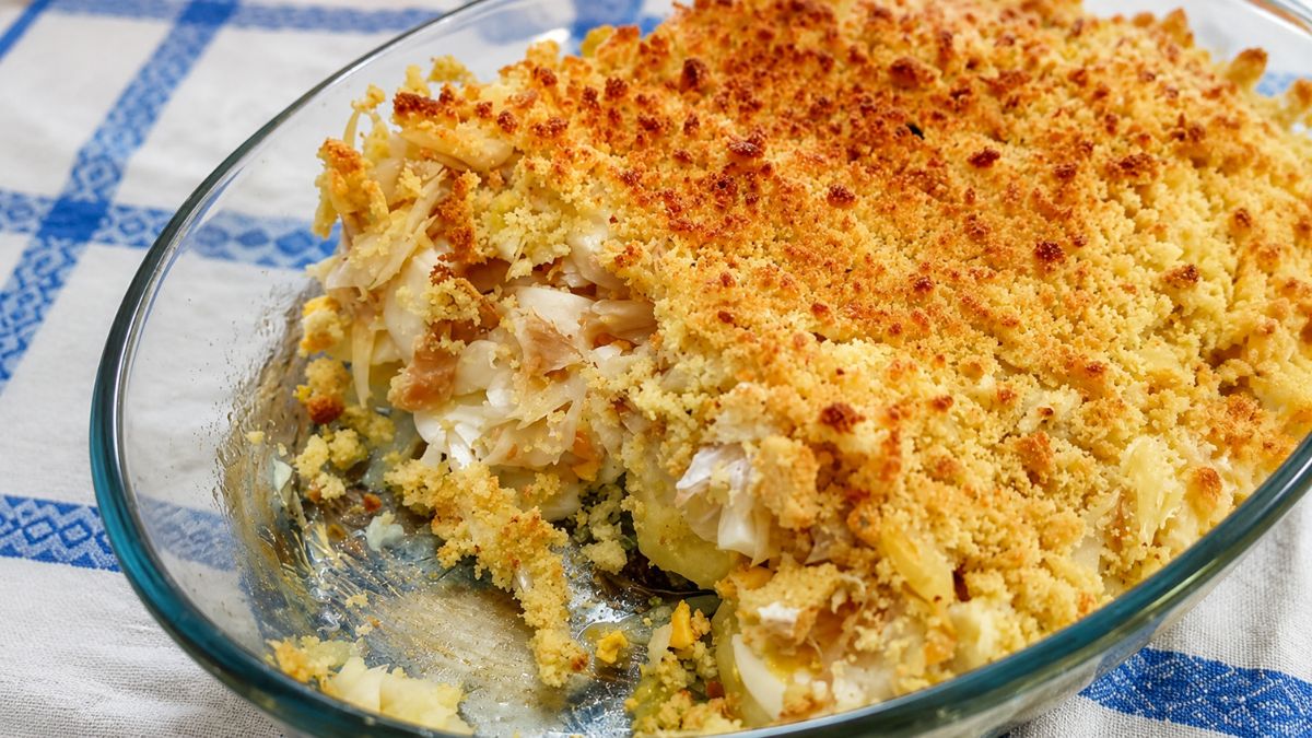 Uma travessa de vidro transparente exibe bacalhau em lascas suculentas, batatas e ovos, tudo coberto por uma generosa camada dourada e crocante de broa de milho gratinada, sobre uma toalha de mesa branca e azul.