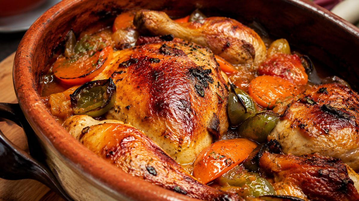 Uma caçarola de barro rústica cheia de frango do campo assado dourado com pele crocante, cozinhas com legumes mistos, incluindo cenouras, tomates e pimentões em um molho rico.