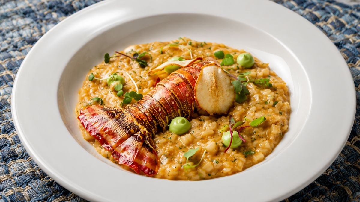 Fotografia gastronômica em plano fechado de um sofisticado risoto de lagosta cremoso servido em prato branco, finalizado com uma cauda de lagosta inteira grelhada, brotos verdes frescos e pequenas esferas de purê sobre toalha de tecido azul texturizado.