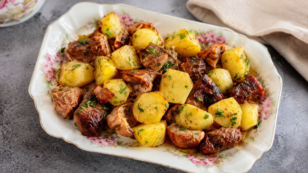 Prato de porcelana decorado com flores servindo rojões no forno, destacando cubos de carne de porco dourados e batatas assadas salpicadas com coentros frescos picados.