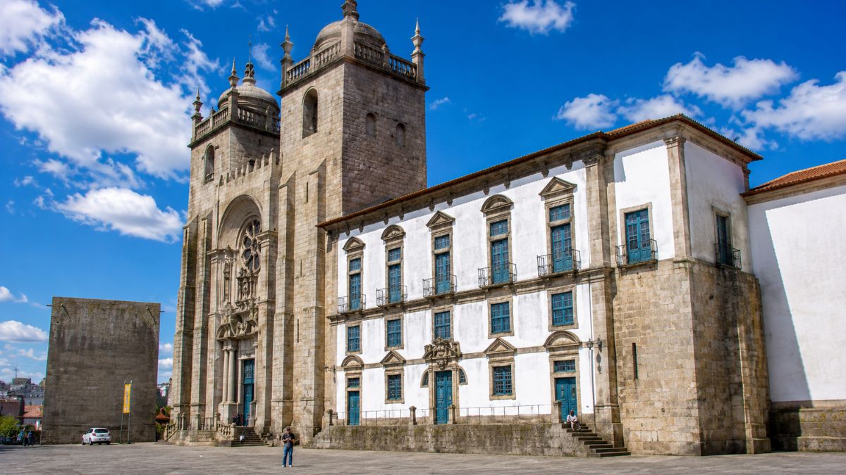 Grande praça seca do Terreiro da Sé exibindo o imponente edifício do Paço Episcopal com janelas azuis ao lado das torres da Sé Catedral do Porto sob céu azul.