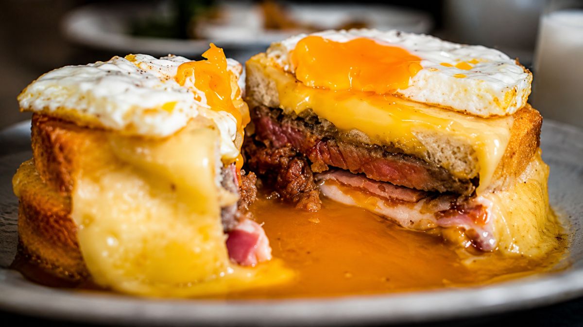 Sanduíche francesinha do Porto cortado ao meio com camadas de carne, queijo derretido, ovo frito e molho picante no prato.