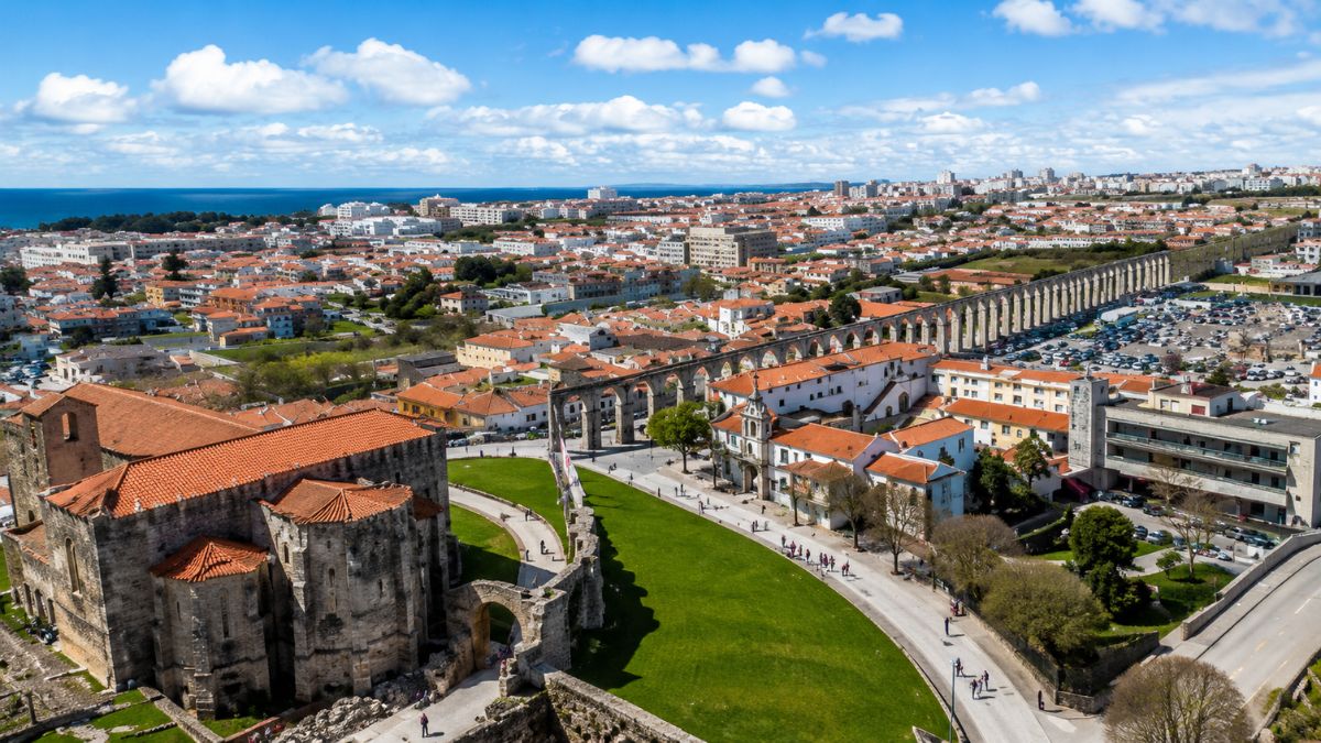 Fotografia aérea mostrando em primeiro plano a estrutura de pedra do Mosteiro de Santa Clara ao lado do extenso Aqueduto de Vila do Conde com seus arcos antigos atravessando a cidade sob céu azul com nuvens.