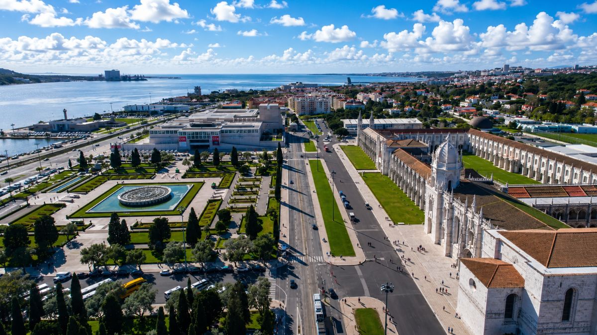 Fotografia aérea mostrando o Mosteiro dos Jerónimos à direita com sua arquitetura manuelina, a Praça do Império com jardim geométrico e fonte central à esquerda, e o Rio Tejo ao fundo sob céu azul com nuvens em Lisboa Portugal.