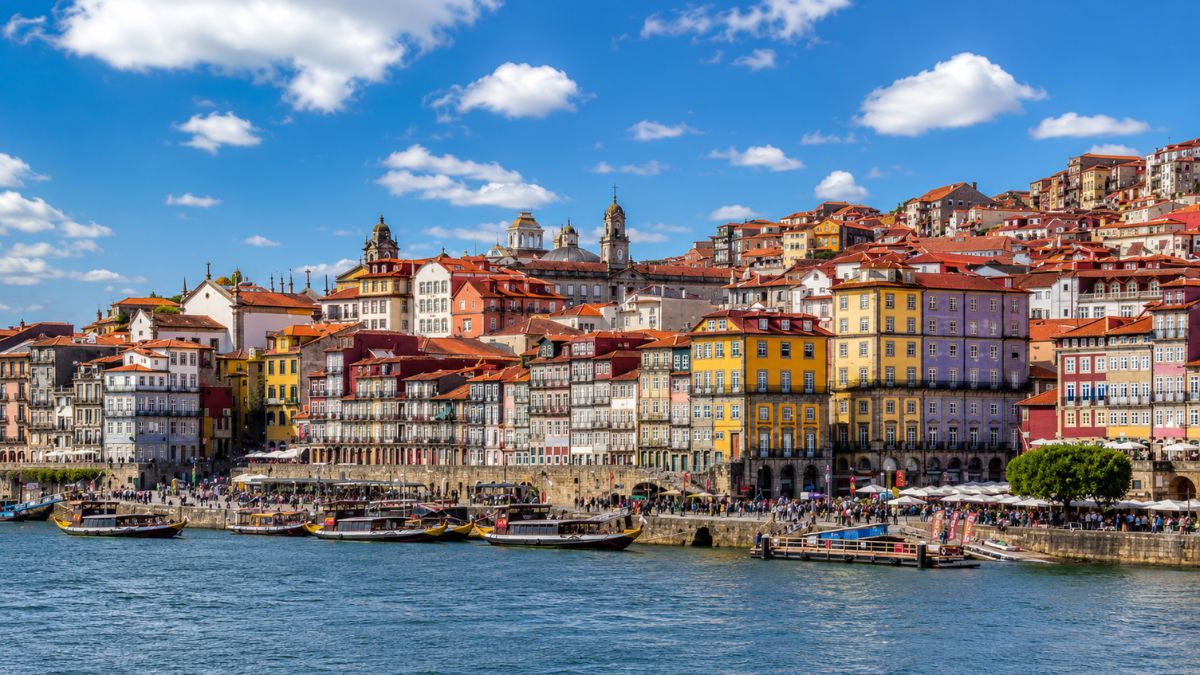 Casas coloridas da Ribeira na encosta do rio Douro com barcos rabelos ancorados sob céu azul com nuvens no Porto.