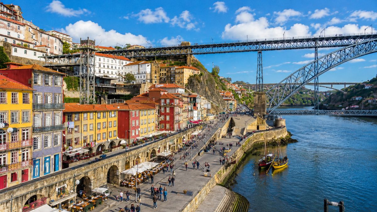 Casas coloridas da Ribeira à beira do Rio Douro com a imponente Ponte Luís I ao fundo em um dia ensolarado no Porto.
