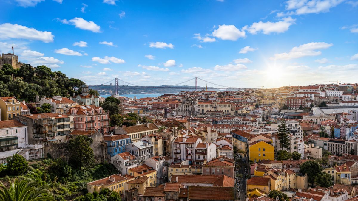 Fotografia aérea de Lisboa mostrando o casario histórico com telhados vermelhos sob céu azul com nuvens, destacando a Ponte 25 de Abril ao fundo sob a luz do sol.